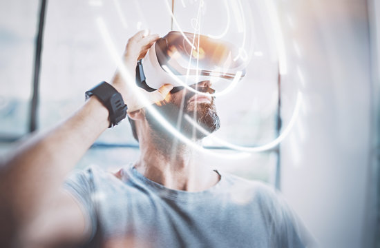 Concept of digital screen,connection and interfaces.Attractive bearded man enjoyingvirtual reality glasses in modern loft studio.Smartphone using with VR goggles headset.Flare effect,blurred.