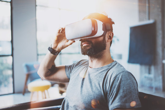 Attractive bearded man wearing virtual reality glasses in modern interior coworking studio.Hipster using smartphone with VR goggles headset.Horizontal,film effect,flare, blurred background.