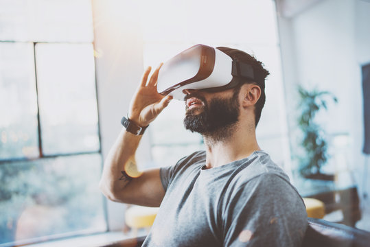 Attractive bearded man using virtual reality glasses in modern interior coworking studio.Hipster enjoying smartphone with VR goggles headset.Horizontal,film effect,flare, blurred background.
