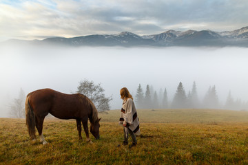 Obraz premium Beautiful girl with horses at mountain peak above clouds and fog. Girl with an horse playing together at the farm. Young woman take care of her horse. Early morning.Misty mountain.