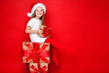 Happy smiling girl in Christmas cap with gift wrapped in craft paper and red ribbons sitting on the gifts on red background. Christmas. Happy family.