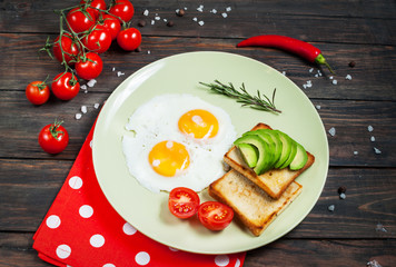 Pan of fried eggs and cherry-tomatoes with bread, avocado on dark wood table