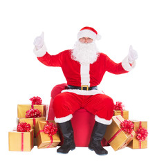 Santa Claus sitting at big red Christmas sack and golden presents, gifts and surprises around isolated on white background, thumbs up gesturing. Happy New Year or Merry Xmas holiday concept