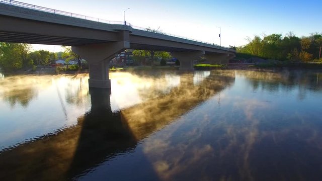 Rays of sunshine illuminate steaming river at sunrise, as swallows dive down from under bridge.