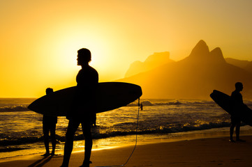 Silhouettes of surfers holding their surfboards on the background of golden sunset on Ipanema Beach and Two Brothers, Dois Irmaos Mountain, Rio de Janeiro, Brazil 