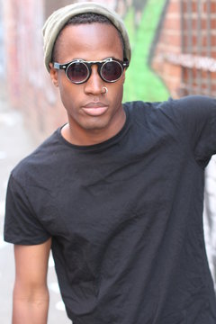 Fashion african man wearing a sunglasses, beanie, piercing and black tee over urban background in city alley