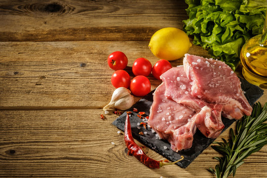 Raw pork steaks on stone board with herbs, tomatoes, garlic and