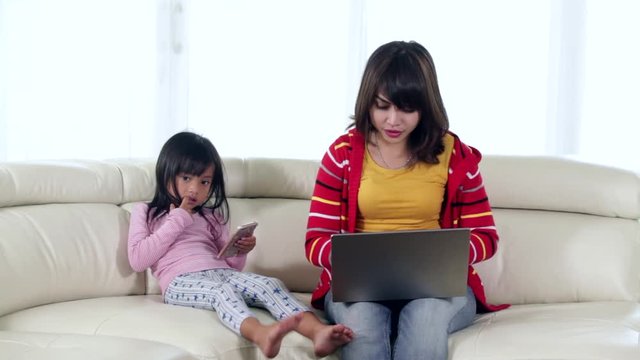 Little girl playing games with a smartphone while her mother working with a laptop computer on the sofa at home