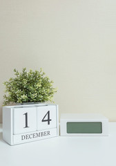 White wooden calendar with black 14 december word with clock and plant on white wood desk and cream wallpaper textured background , selective focus at the calendar