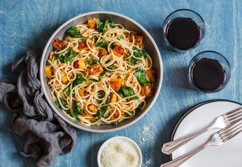 Baked pumpkin and spinach spaghetti in a frying pan and two glasses of red wine on wooden background, top view. Delicious lunch in a mediterranean style. Flat lay