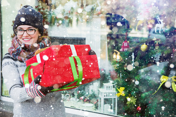 Obraz premium A girl stands at the storefront, on a city street. Shopping, she buys gifts. Woman holding a red box gifts. Background seasonal winter holidays.