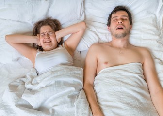 Young woman is lying with her husband in bed. Man is snoring too loud. Woman is covering her ears.