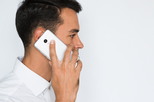 Sideways Portrait Of Handsome Young Smile Man In White Shirt Using Mobile Phone, Looking At The Camera With Serious And Concentrated Expression. Businessman Sale By Smart Phone On Gray Background.