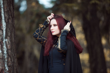 Young beautiful and mysterious woman in woods, in black cloak with hood, image of forest elf or...