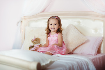 Fototapeta na wymiar Very nice charming little girl blonde in pink dress with teddy bear sitting on a child's bed and laughs loudly in bright interior of the house