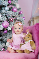 charming little girl with blue eyes sitting in a pink armchair teddy bear on background decorated Christmas tree