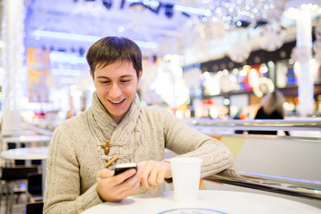 Handsome young man in white sweater using a smart phone, drinking coffee and smiling while resting in cafe after doing shopping in the mall. Christmas time