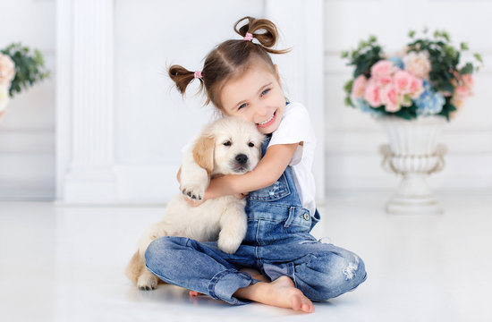 Little girl,hair tied with pink bands into two tails,dressed in a white t-shirt and blue denim overalls is playing at home,sitting alone on the bed with their favorite dog breed Golden Retriever