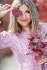 Autumn portrait of beautiful young woman with long blonde hair and grey eyes,dressed in a pink knitted sweater,spending time outdoors in the autumn Park among the trees and shrubs with red foliage
