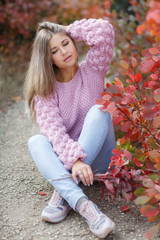Autumn portrait of beautiful young woman with long blonde hair and grey eyes,dressed in a pink knitted sweater,spending time outdoors in the autumn Park among the trees and shrubs with red foliage