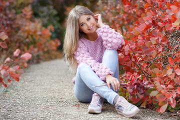 Autumn portrait of beautiful young woman with long blonde hair and grey eyes,dressed in a pink knitted sweater,spending time outdoors in the autumn Park among the trees and shrubs with red foliage