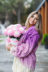 Beautiful woman with long straight blond hair and grey eyes, dressed in a beautiful knitted coat,holding a large bouquet of pink flowers autumn portrait on one of the streets of the city