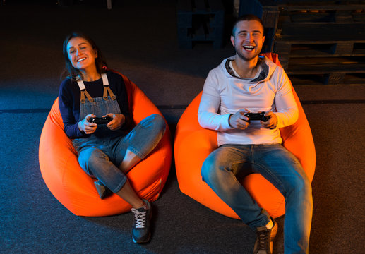 Two young gamer sitting on poufs and playing video games togethe