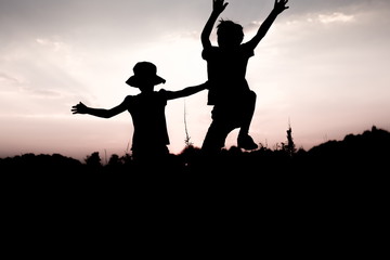 Silhouettes of kids jumping off a cliff at sunset. Little boy and girl jump raising hands up high. Brother and sister having fun in summer. Friendship, freedom concept.
