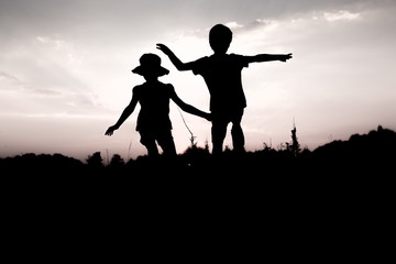 Silhouettes of kids jumping off a cliff at sunset. Little boy and girl jump raising hands up high. Brother and sister having fun in summer. Friendship, freedom concept. Twins on vacation