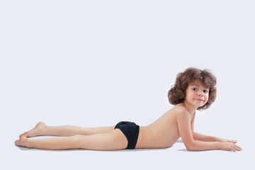 Cute curly-haired boy in shorts lying on the floor and looking into the camera. Grey background.