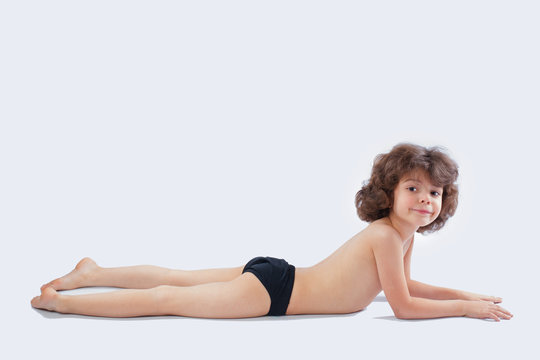 Cute curly-haired boy in shorts lying on the floor and looking into the camera. Grey background.