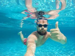 Beard man with glasses diving in a blue clean water with thumb u