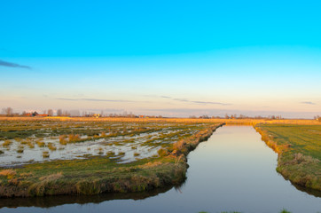 A flooded and marshy field of grass by a canal against a blue sky background