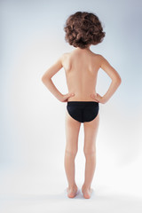 Curly boy in swimming trunks standing in full length with his back to the camera. Gray background.