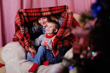 Mom and son playing in the house under a blanket on the background of Christmas tree.waiting for the Santa Claus