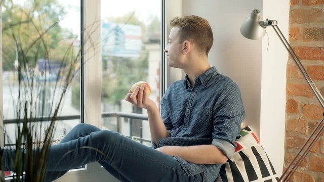 Man looking thoughtful while sitting by the window and eating apple
