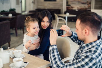 family, parenthood, technology, people concept - happy father taking photo of his little daughter and wife by smartphone having dinner at restaurant