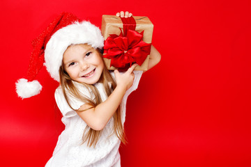 Happy smiling girl in Christmas cap with gift wrapped in craft paper and red ribbons on red background. Christmas. Happy family.