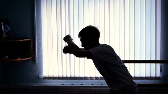 Close up of silhouette of a young hip-hop dancer in black trousers and white shirt performing elements of breakdance against the window in the shadowed room.