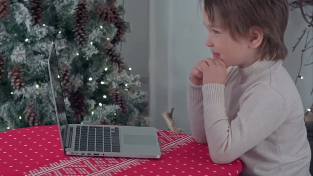 Boy using his laptop sitting at the table near the Christmas tree