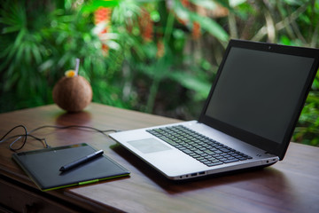 Tropical freelance: graphic tablet with pen and laptop on wooden table on background of green palms and cocounat.