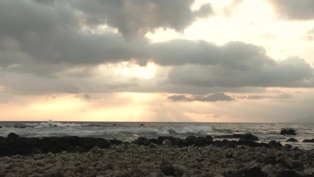 Time lapse from Maui, Hawaii at beach during sunset with sun dropping behind clouds and waves crashing on shore.