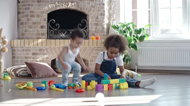 Little African girl sitting on the floor in the living room and building something with toy blocks while brother standing behind her