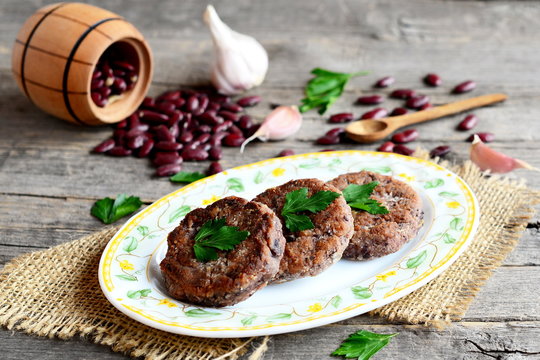 Burgers made from boiled and mashed red beans on a plate. Scattered uncooked red beans, garlic, fresh parsley, small spoon and decorative barrel on old wooden background. Vegetarian burgers. Closeup