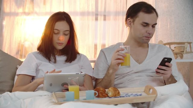 Young woman sitting in bed looking at tablet and drinking juice from breakfast tray alongside her husband with mobile phone