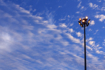 Large tall high outdoor stadium halogen spotlight with lamp light turned on and sky clouds in twilight.
