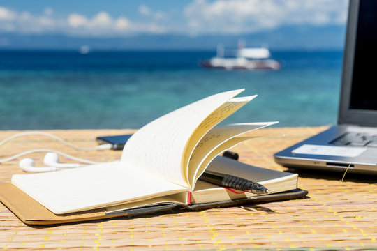 Opened empty notepad is on the table with phone and headphones at the tropical sea background