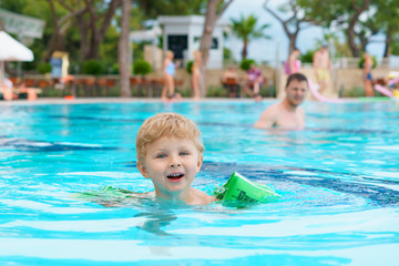 Fototapeta na wymiar Little blond kid learning to swim in the resort pool. Dressed inflatable armbands. Watching instructor on a blurred background.