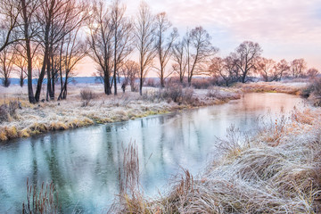silhouettes of trees on the river bank. Yellow dry grass on the shore covered with the first frost, the beautiful sky with clouds. Beautiful landscape in late autumn,  in winter early morning.
