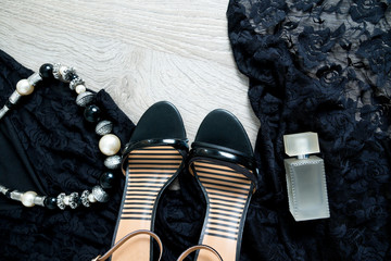 Women outfit. Black lace dress, shoe sandals, necklace and perfume on grey wooden background. Top...